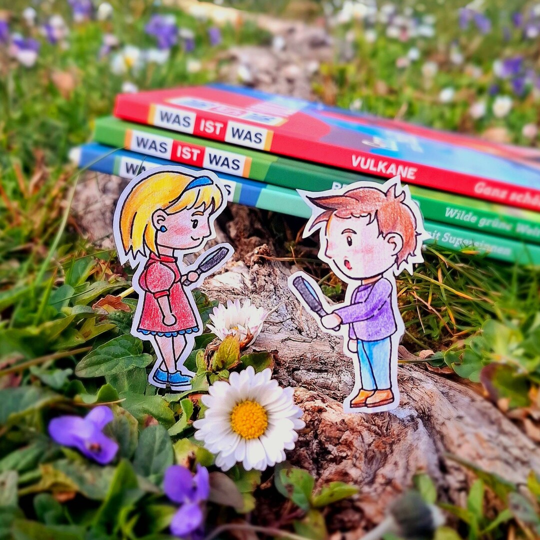 Drei "Was ist was"-B&uuml;cher liegen im gras, man sieht die Buchr&uuml;cken. Davor stehen zwei Comic-Personen, die auf Papier gemalt und dann ausgeschnitten wurden. Sie halten Lupen in der Hand. Im Vordergrund ist ein G&auml;nsebl&uuml;mchen zu sehen.