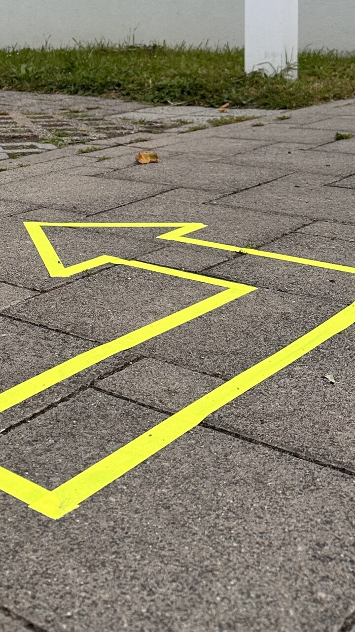 Drau&szlig;en wurde auf einem Weg auf dem Campusgel&auml;nde ein Pfeil mit neongelbem Klebeband geklebt. Er dient als Wegweiser.